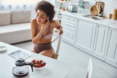Mutlu Afro-Amerikalı kadın iç çamaşırlarıyla kulaklıklara dokunuyor ve sabahları kahve içiyor.