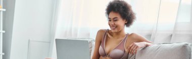 Mutlu Afro-Amerikalı kadın iç çamaşırlarıyla kanepede oturuyor ve dizüstü bilgisayarda film izliyor.