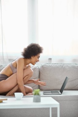 happy african american woman in lingerie sitting on couch and using laptop at home, entertainment clipart