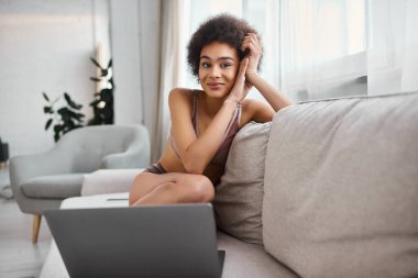 İç çamaşırlı Afrikalı Amerikalı bir kadın dizüstü bilgisayarın yanındaki koltukta oturmuş kameraya bakıyordu.