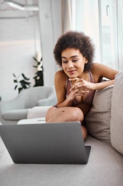 Afro-Amerikan kadın iç çamaşırlarıyla kanepede oturuyor ve dizüstü bilgisayarda film izlerken gülümsüyor.