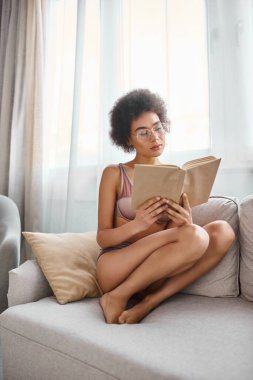 Rahat ve kıvırcık saçlı Afrikalı Amerikalı kadın rahat bir kanepede iç çamaşırlarıyla kitap okuyor.