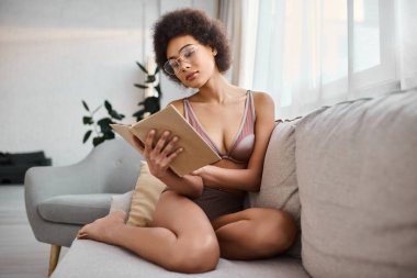 Rahat ve kıvırcık saçlı Afrikalı Amerikalı kadın rahat koltukta iç çamaşırlarıyla kitap okuyor.