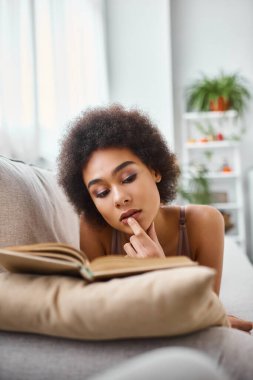 concentrated and young african american woman reading a book in lingerie on a comfy sofa clipart