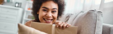 cheerful and young african american woman holding a book and lying on couch at home, banner clipart