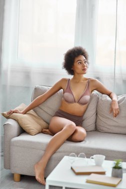 dreamy young african american woman with curly hair lounging in lingerie on a sofa at home clipart