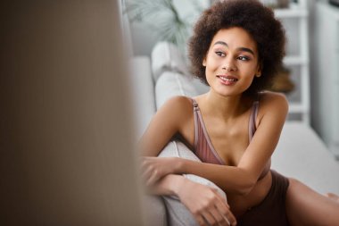 Mutlu Afro-Amerikalı kadın, kıvırcık saçlı, iç çamaşırlarıyla kanepede dinleniyor, gülümsüyor ve başka tarafa bakıyor.
