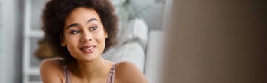 Mutlu Afro-Amerikalı kadın, kıvırcık saçlı, iç çamaşırlarıyla kanepede dinleniyor, gülümsüyor.