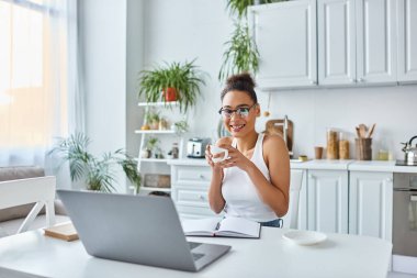 positive african american woman in glasses holding cup of coffee near laptop on desk, remote work clipart