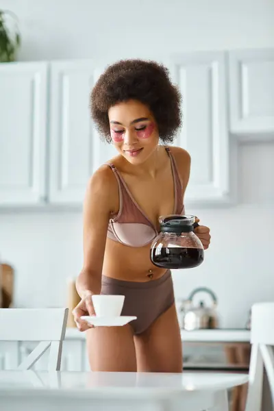 Radiant curly african american woman with eye patches holding coffee pot and cup in kitchen