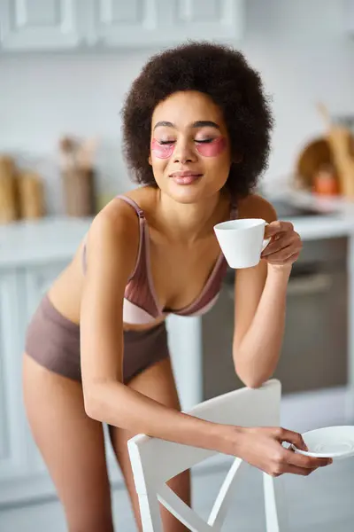 joyful african american woman with eye patches standing in underwear and enjoying morning coffee