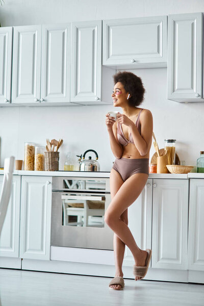 Serene moment of happy african american woman in earphones and lingerie sipping morning coffee