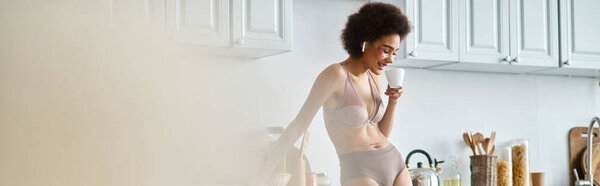 banner of happy african american woman in earphones and lingerie sipping morning coffee in kitchen