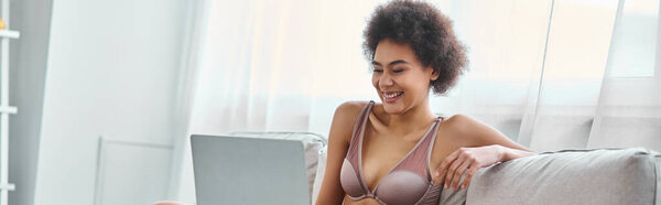 happy african american woman in lingerie sitting on couch and watching movie on laptop, banner