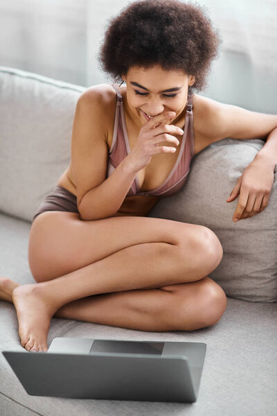 african american woman in lingerie sitting on couch and laughing while watching movie on laptop
