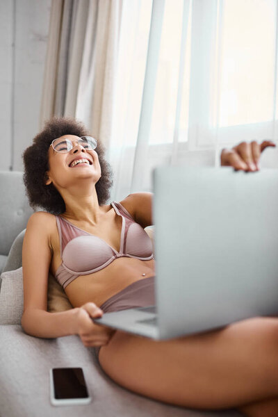 african american woman in lingerie with glasses sitting on couch and laughing from comedy movie