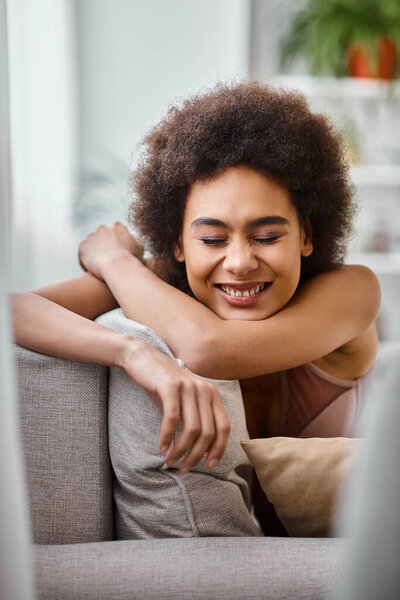 happy african american woman with curly hair relaxing on couch in lingerie, smiling with closed eyes