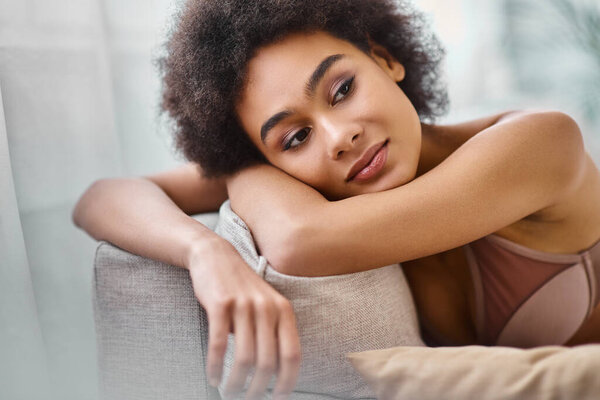 Dreamy african american woman with curly hair relaxing on couch in lingerie, lost in thoughts