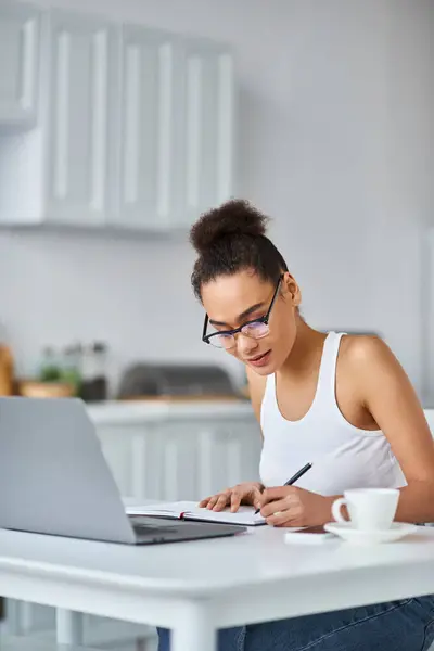 带着眼镜 卷曲的非洲裔美国女人 在家里的笔记本电脑上做着远程工作 做着笔记 — 图库照片