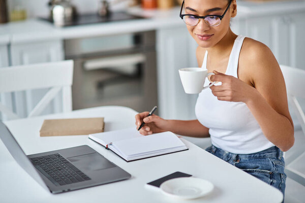 curly african american woman in glasses working from home remotely on her laptop, sipping coffee