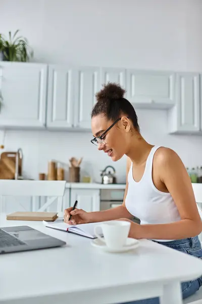 stock image happy african american woman in glasses taking notes near laptop and cup of coffee, remote work