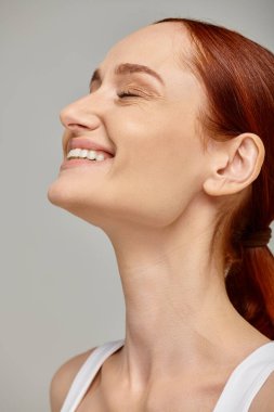 Mutlu kızıl saçlı, gözleri kapalı, gri arka planda neşeli ve sağlıklı bir gülümseme saçan bir kadın.
