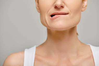 cropped view of pensive woman in white tank top biting lip, embodying concern on grey background clipart