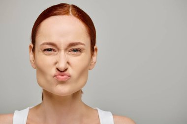 redhead young woman in white tank top frowning and grimacing on grey background, concerned face clipart