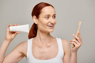 amazed woman with red hair holding toothpaste and toothbrush and smiling at camera on grey backdrop clipart