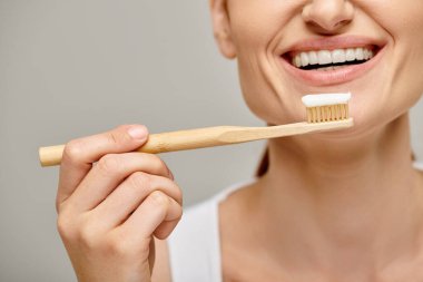 cropped view of joyful woman holding toothbrush with toothpaste and smiling at camera on grey clipart