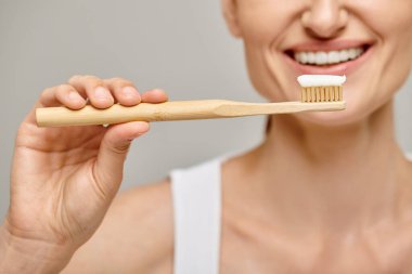 cropped view of cheerful woman holding toothbrush with toothpaste and smiling at camera on grey clipart