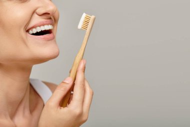 cropped shot of joyful woman holding toothbrush with toothpaste and smiling on grey backdrop clipart