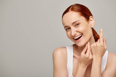 excited redhead model holding dental floss and smiling on grey background,  promoting oral hygiene clipart