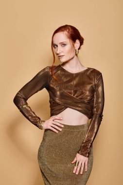 30 'lu yaşlardaki güzel kızıl saçlı kadın altın rengi elbiseler ve bej arka planda aksesuarlarla poz veriyor.