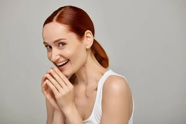 astonished and redhead woman in white tank top laughing a grey background, radiant smile