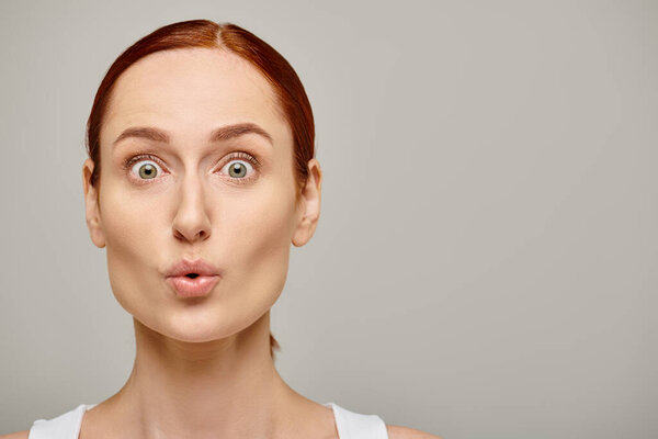 surprised young woman in white tank top looking at camera with wide eyes on grey background, wow