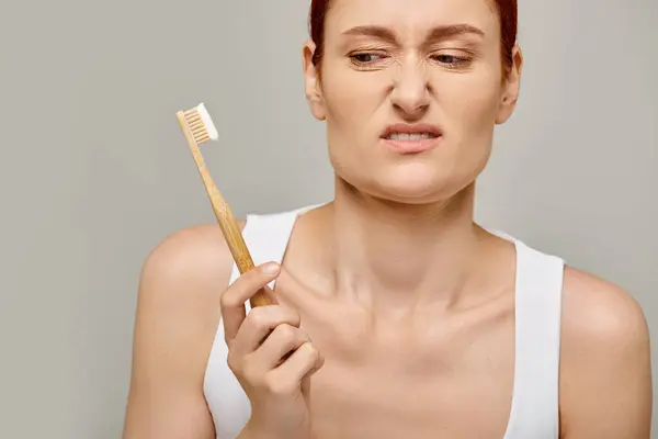 stock image displeased woman with red hair looking at bamboo toothbrush with toothpaste on grey backdrop, dental