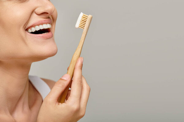 cropped shot of joyful woman holding toothbrush with toothpaste and smiling on grey backdrop