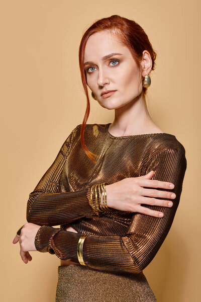 redhead woman in her 30s posing in glamorous attire with golden accessories on beige backdrop