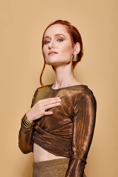 redhead woman in her 30s posing in glamorous attire and golden accessories on beige backdrop