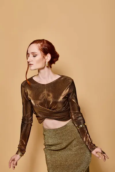 stock image redhead model in her 30s posing in glamorous attire and golden accessories on beige backdrop