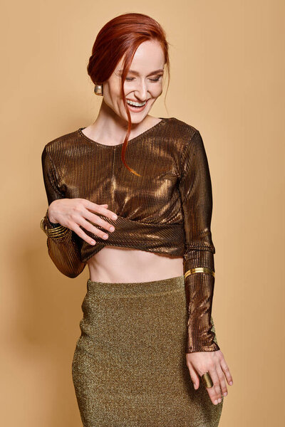 happy redhead woman in her 30s posing in trendy attire and golden accessories on beige backdrop