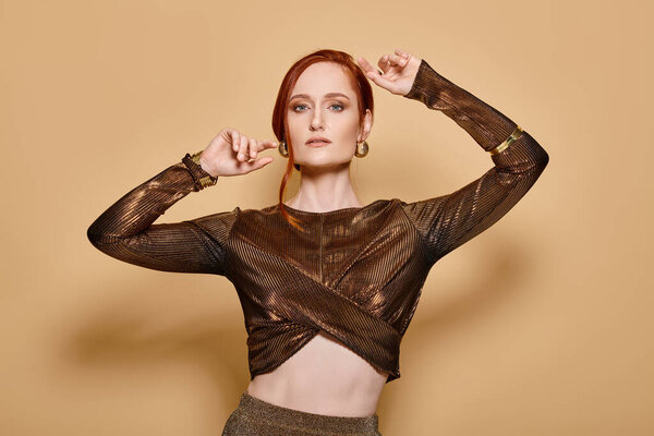 redhead woman in her 30s posing in stylish attire and golden accessories with raised hands on beige
