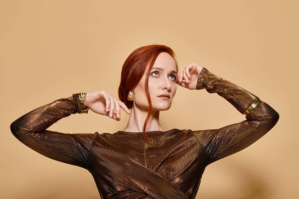 dreamy woman in her 30s posing in stylish attire and golden accessories with raised hands on beige