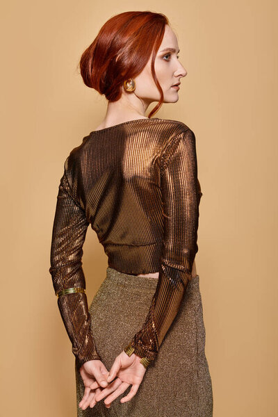 redhead woman in her 30s posing in trendy attire and golden accessories with hands behind back