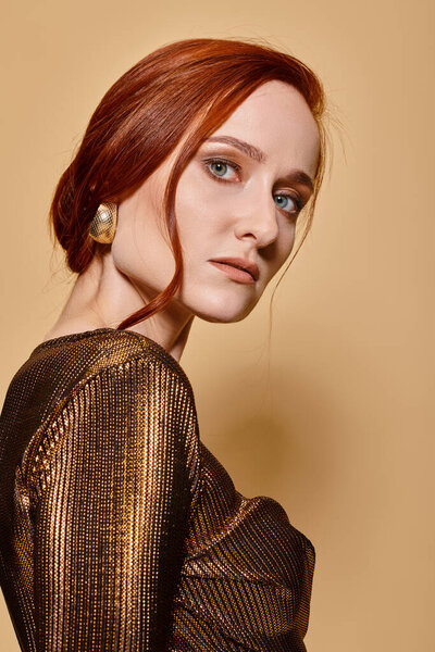 portrait of alluring redhead woman in 30s posing in trendy attire and golden accessories on beige
