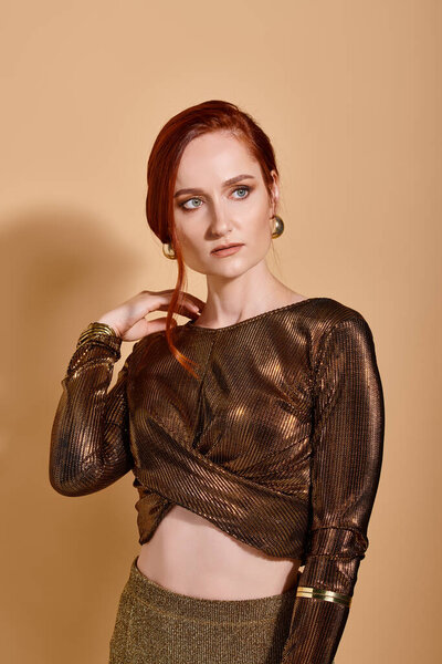 dreamy redhead woman in 30s posing in stylish outfit and golden accessories on beige backdrop
