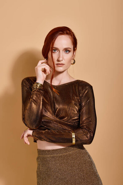 thoughtful redhead woman in 30s posing in stylish outfit and golden accessories on beige backdrop