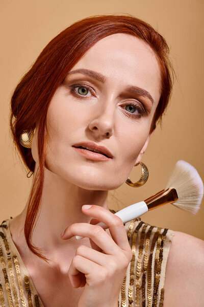 Redhead woman in gold sequin dress holding makeup brush on beige background, beauty gaze