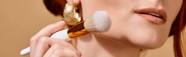 cropped redhead woman in gold earring holding makeup brush for liquid face foundation, banner